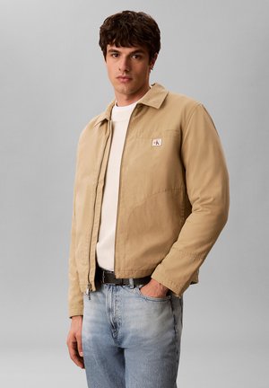 Man draagt een beige jas met rits over een witte trui en lichtblauwe spijkerbroek, staand met één hand in de zak tegen een grijze achtergrond.