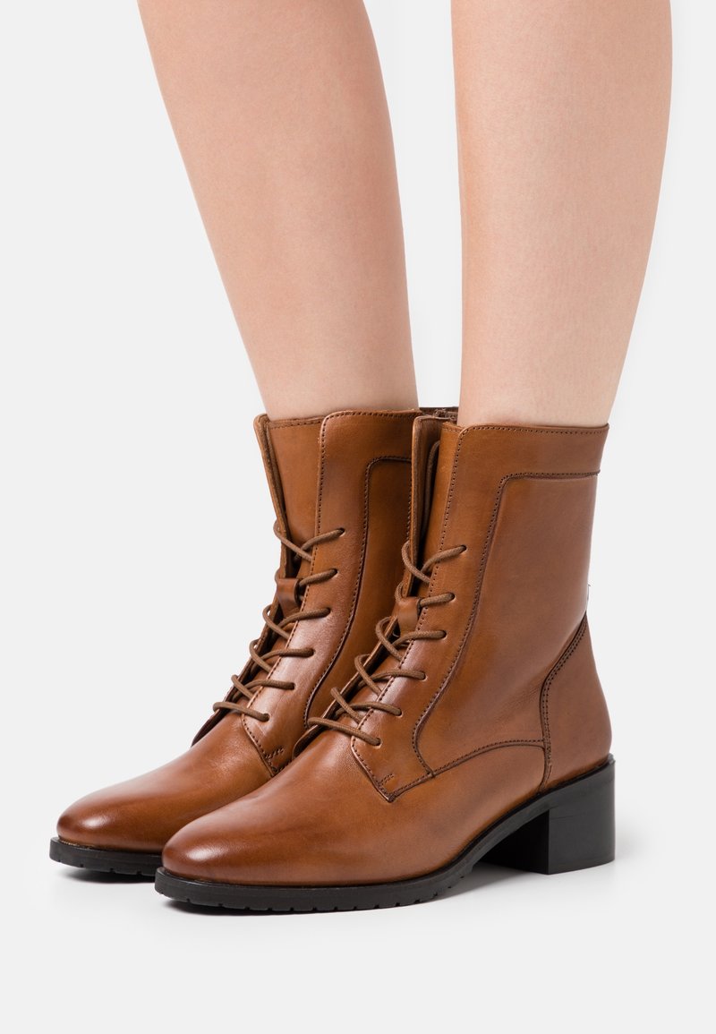 Bottines en cuir marron avec un design à lacets, un talon carré et des détails de couture le long des côtés et de l'orteil. Texture lisse, accents minimaux.