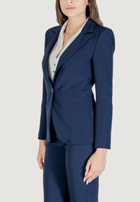Blazer su misura di colore blu navy con rever a punte, due tasche frontali e chiusura con un solo bottone. Tessuto liscio con una vestibilità strutturata.