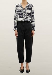 Kvinna i svarta högmidjade avsmalnande jeans, blus med svart och vit grafisk tryck samt svarta peka-tå klackar med ankelsnören.