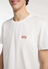 Camiseta de algodón blanca con un pequeño diseño de furgoneta bordado en naranja en la parte izquierda del pecho. Mangas cortas y cuello redondo.