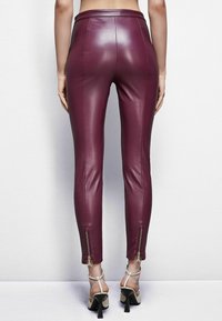 Pantaloni in pelle bordeaux con un design aderente, cuciture verticali e cerniere alla caviglia con hardware decorativo. Indossati con tacchi alti con cinturini.