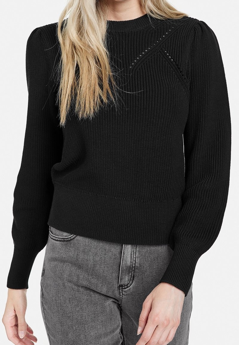 Femme portant un pull noir en tricot avec des épaules bouffantes et un jean gris taille haute, debout devant un fond uni.