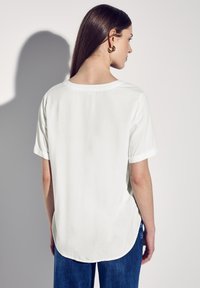 Blusa bianca a maniche corte con scollatura rotonda, texture morbida e orlo leggermente curvo. Abbinata a pantaloni in denim blu.