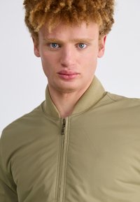 Olivovo zelená bomber bunda so zipom, rebrovaným golierom a hladkým, mäkkým povrchom. Jednoduchý dizajn bez viditeľných vzorov alebo hardvéru.