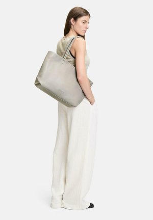 Graue Leder-Tote-Bag mit glatter Textur und breiter Form. Verfügt über lange Träger und ein minimalistischem Design. Das Modell trägt hellfarbige Kleidung.
