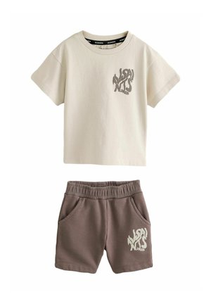 Bambino piccolo in piedi di spalle, che indossa una maglietta crema con testo nero, pantaloncini marroni, calzini bianchi e scarpe da ginnastica bianche su sfondo grigio.