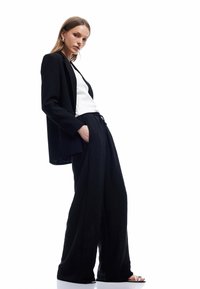 Zwarte blazer met een op maat gemaakte pasvorm, gecombineerd met losse, getextureerde zwarte broek. Witte top eronder en minimalistische sandalen.