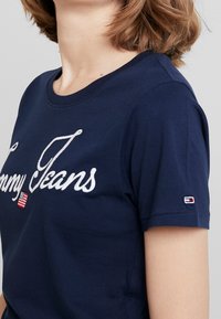 Tmavě modré bavlněné tričko s krátkými rukávy, s bílým vyšitým nápisem "Tommy Jeans" a malým logem na rukávu.