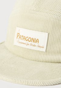 Μπεζ καπέλο κορδούρας με ραμμένη λευκή ετικέτα που απεικονίζει την επιγραφή «Patagonia Equipment for Water People» σε χρυσά γράμματα.