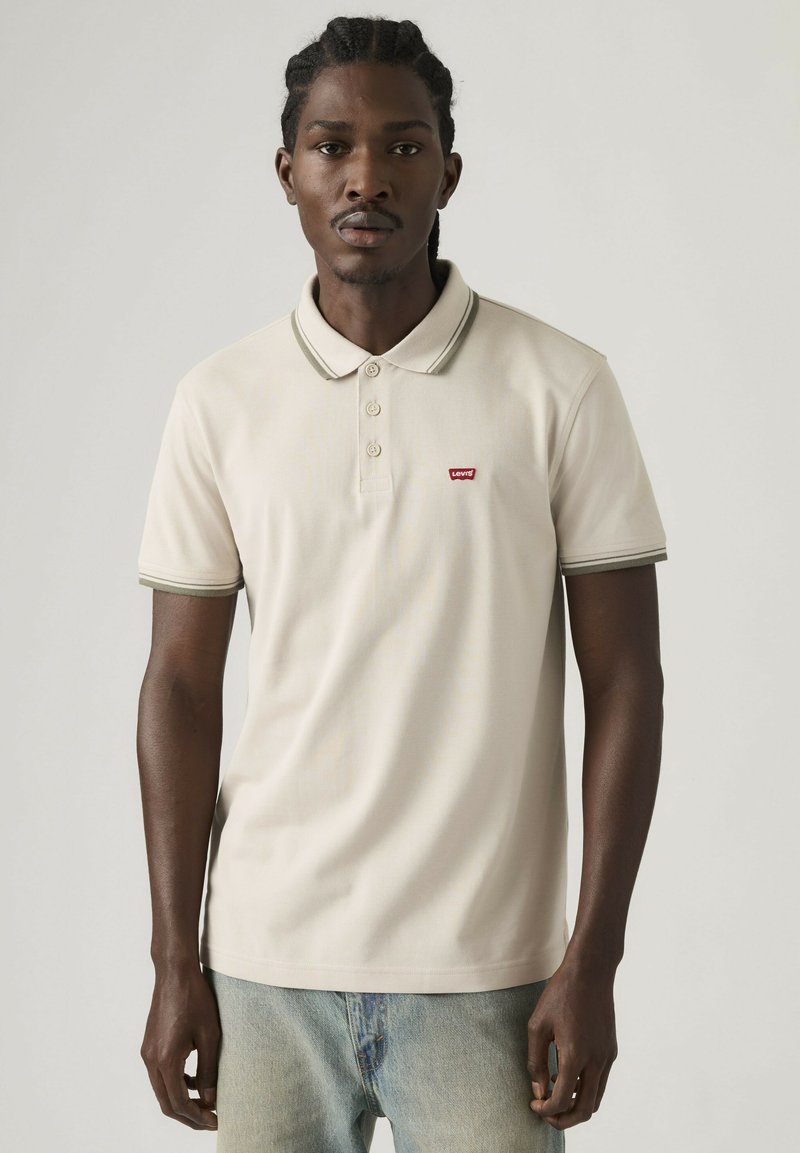 Homme aux cheveux tressés portant un polo Levi's beige clair et un jean bleu clair, debout devant un fond gris uni.