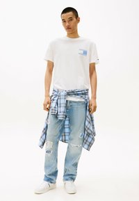 Hvid t-shirt med blå mønstret lomme, lyseblå ternet skjorte bundet om livet, slidte lyseblå jeans og hvide sneakers.