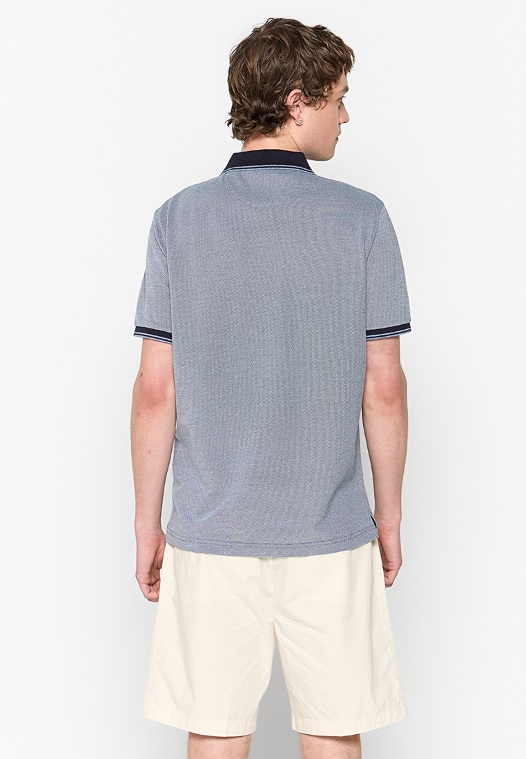 Kortebloem polo shirt in lichtblauw, met een getextureerd patroon, marinekleurige kraag en boorden, en een rechte zoom. Gekombineerd met lichte korte broek.