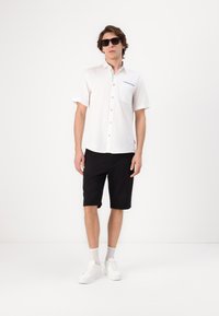 Camisa blanca de manga corta con un bolsillo en el pecho, combinada con pantalones cortos negros hasta la rodilla y zapatillas blancas. El conjunto presenta elementos de diseño mínimos.