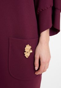 Hand, die nahe einer burgunderfarbenen Kleidertasche ruht, geschmückt mit einem abstrakten goldenen Brosche.