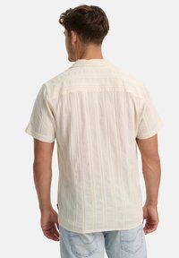 Homme portant une chemise texturée crème à manches courtes et un jean bleu clair, debout de dos face à la caméra sur un fond blanc.