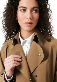Beige trenchcoat med stor krave, trykknapper og en glat tekstur, lagd over en hvid skjorte med krave.