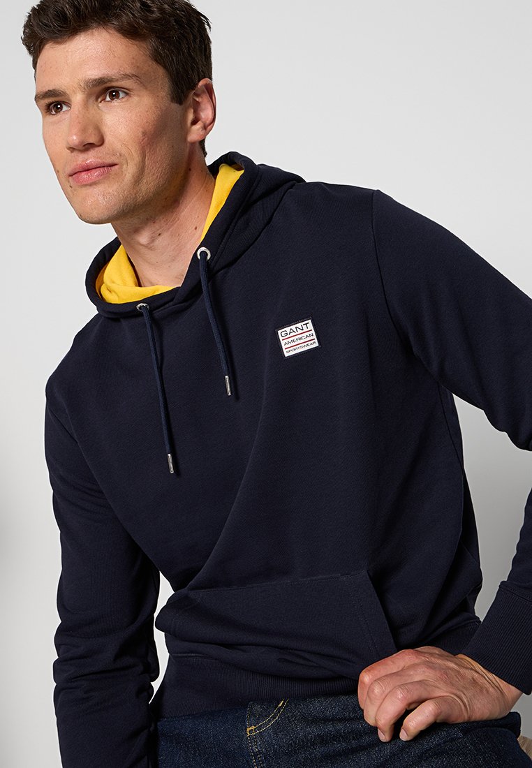 Sweat à capuche bleu marine avec doublure jaune, cordons de serrage, poche avant et patch logo sur la poitrine. Texture douce et design décontracté.