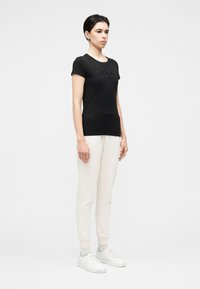 Melnas kokvilnas T-krekls ar īsām piedurknēm un reljefainu detalizāciju krūšu daļā, kombinēts ar krēmkrāsas sporta biksēm un baltām sporta kurpēm.