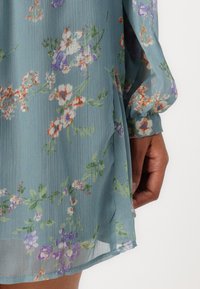 Lettvekts, gjennomsiktig kjole i et mykt blått stoff med blomsterdesig i oransje, lilla og grønt. Har luftige ermer og en sidesplitt.