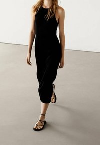 Schwarzes geripptes Kleid mit tailliertem Oberteil und mittellangem Saum. Kombiniert mit schwarzen flachen Sandalen mit überkreuzten Riemen.