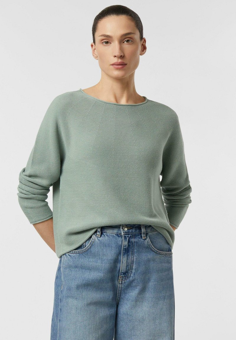 Femme aux cheveux foncés portant un pull en maille vert sauge et un jean taille haute bleu clair, debout devant un fond clair uni.