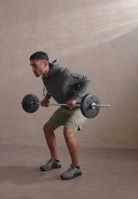 Svorio kilnojimo pratimas, kuriame matomas asmuo pilkoje gobtuvinėje striukėje ir smėlio spalvos šortuose, laikantis štangos su juodais svoriais sporto salėje.