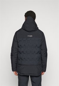 Crna puf jakna s kapuljačom, s prešanim uzorkom i Columbia logotipom na gornjem dijelu leđa. Glatka tekstura, prilagodljivi rub.