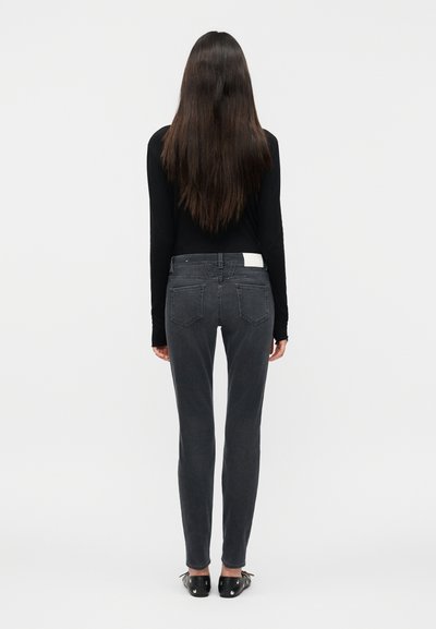 Femme aux longs cheveux bruns portant un haut noir à manches longues, un jean gris foncé ajusté et des chaussures plates noires, debout de dos.