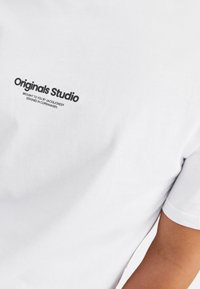 Weißes Baumwoll-T-Shirt mit kurzen Ärmeln, auf der oberen linken Brust mit einem schwarzen bedruckten Logo, das "Originals Studio" liest.