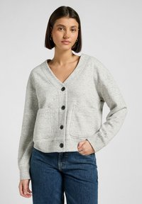 Grå knapplös cardigan med V-ringning, som har två framfickor och ett texturerat stickat mönster. Matcher med blå jeans.