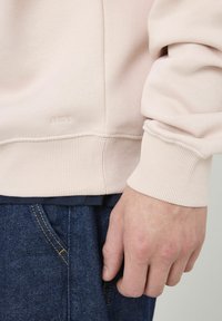 Sweat-shirt rose clair avec des poignets et un ourlet côtelés. Présente une finition texturée et un logo subtil en bas. Associé à un denim foncé.