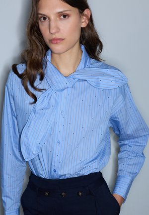 Woman with long brown hair wearing a blue striped blouse with a bow at the collar and dark high-waisted pants against a plain background.