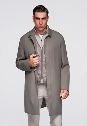 Man draagt een lichtgrijze trenchcoat over een doorgestikte jas met rits en beige broek, staand tegen een effen lichte achtergrond.