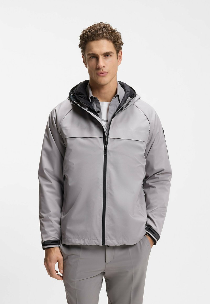 Homme aux cheveux bouclés portant une veste zippée gris clair et un pantalon assorti, debout devant un fond blanc uni.