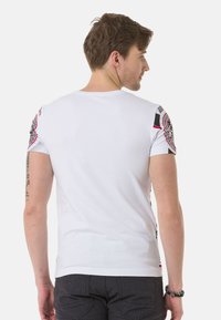 Weißes, eng anliegendes T-Shirt mit schwarzen und roten Schultergrafiken, runder Halsauschnitt und kurzen Ärmeln. Strukturierter Stoff mit glatter Oberfläche.