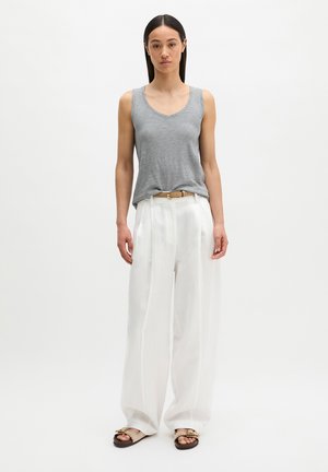 Frau steht vor weißem Hintergrund, trägt ärmelloses graues Oberteil, weiße, hoch taillierte, weit geschnittene Hose, beigen Gürtel und beige Sandalen.