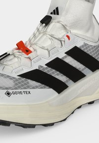 Άσπρο και μαύρο παπούτσι για τρέξιμο Adidas με ύφασμα GORE-TEX, κόκκινες και μαύρες τερματικές ασφάλειες κορδονιών και παχύ μπεζ πέλμα με τραχιά σχεδίαση.