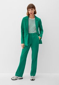 Green blazer over a striped shirt, paired with green trousers. The outfit features a relaxed fit and is complemented by sporty shoes.