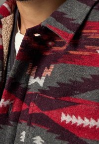 Veste-chemise en laine grise avec des motifs géométriques rouges, bordeaux et blancs ; dotée d'un col en sherpa et d'une poche zippée sur le côté.