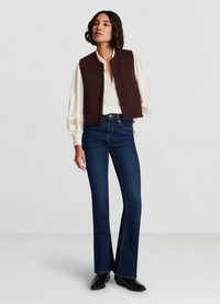 Braune ärmellose Weste über einer cremefarbenen Bluse mit Spitzenbesatz, kombiniert mit dunkelblauen ausgestellten Jeans und schwarzen Stiefeletten, vor hellem Hintergrund.