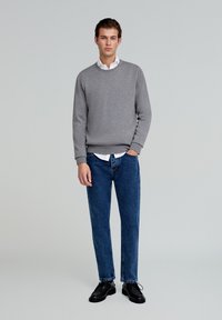 Grijze katoenen trui met ronde hals, gelaagd over een witte overhemdkraag. Gecombineerd met blauwe spijkerbroek en zwarte schoenen.