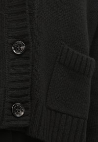 Gros plan d’un cardigan en maille noire avec des poignets côtelés et deux boutons noirs cousus sur la patte de boutonnage.