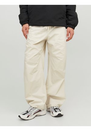 Henkilö, joka pukeutunut löysiin vaaleanbeigeihin housuihin, mustaan takkiin, jossa on joustavat hihansuut, ja valkoisiin, mustiin ja punaisiin urheilukengiin, seisoo yksinkertaisen taustakuvan edessä.