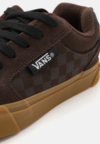 Sneaker Vans marron avec une tige en suède et textile à carreaux, lacets noirs et semelle en caoutchouc couleur gomme. Étiquette logo visible sur le côté.