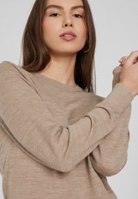 Suéter largo beige de manga larga con cuello redondo. Tejido suave y texturizado con un corte relajado, ligeramente fruncido en las muñecas para un look informal.
