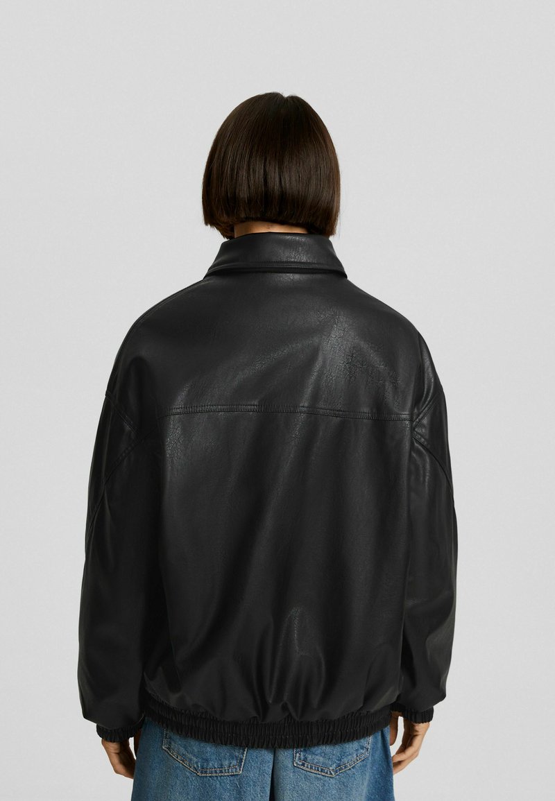 Chaqueta de cuero negro de corte relajado, con gran cuello y puños elásticos. Textura suave con costuras mínimas, visibles desde la parte posterior.