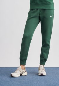 Zaļas kokvilnas maisījuma bikses ar elastīgu vidukļa jostu un aproču pēdām, kopā ar bēšām Nike sporta kurpēm ar baltu logo.