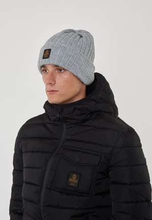 Beanie grigia a maglia con texture a coste e un piccolo logo nero. Indossato con un giubbotto imbottito nero dotato di tasca con zip.