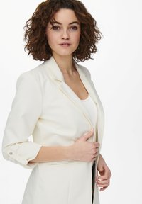 Blazer bianco su misura con chiusura a un bottone, revers a punta e maniche arrotolate. Tessuto liscio e design aderente.
